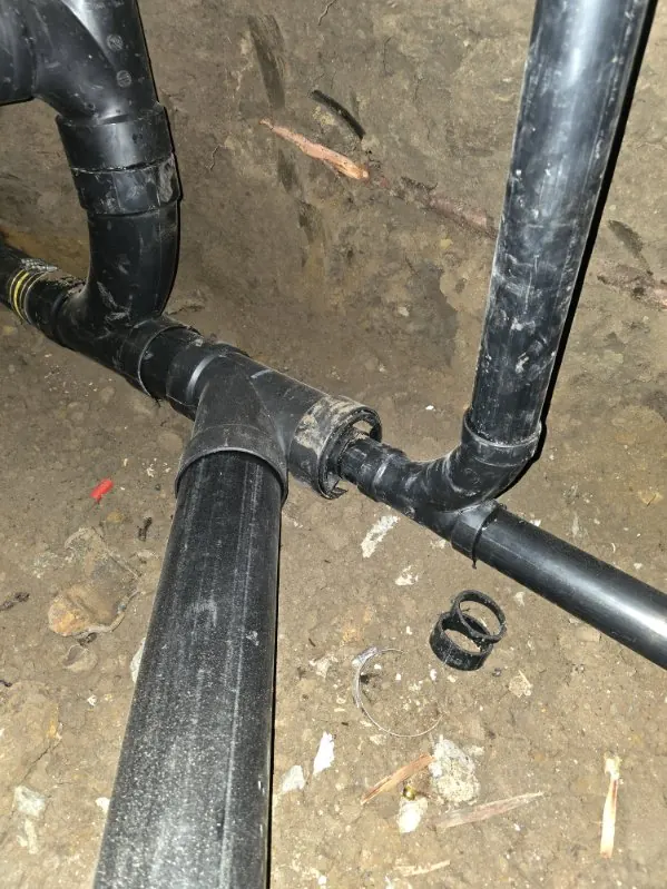 Black ABS drain lines and a wye fitting exposed in soil under a house