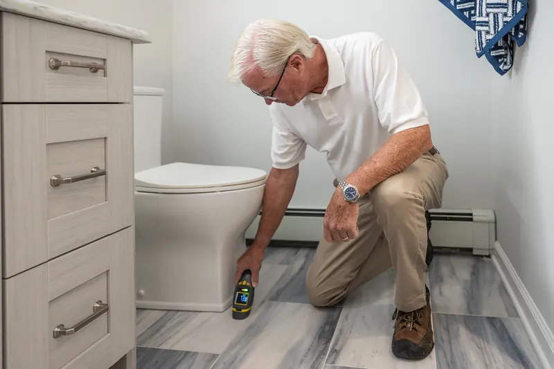 Plumber kneeling by a toilet and testing the floor and baseboard with a moisture meter