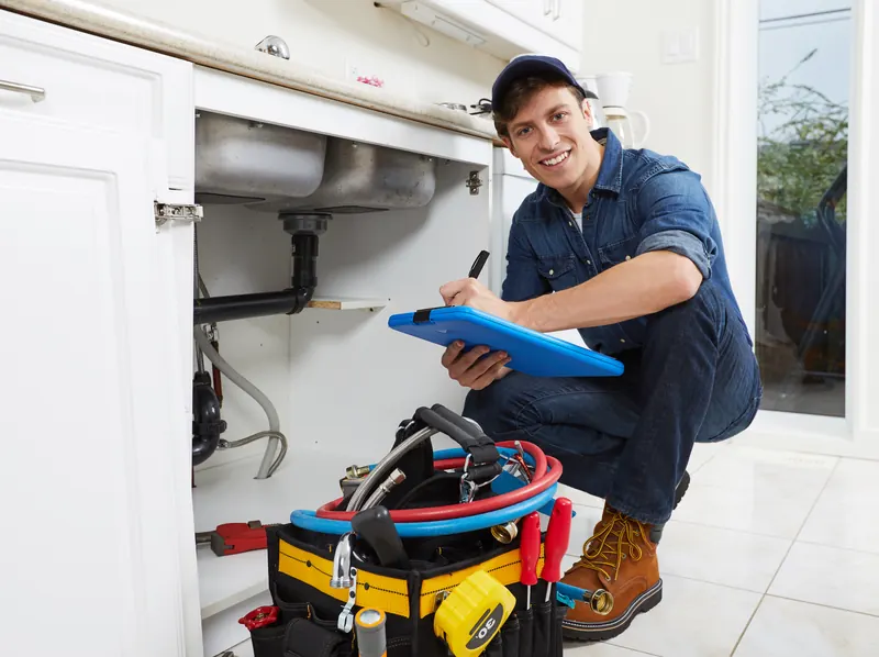 Smiling plumber writing an estimate on a tablet beside an open kitchen sink cabinet